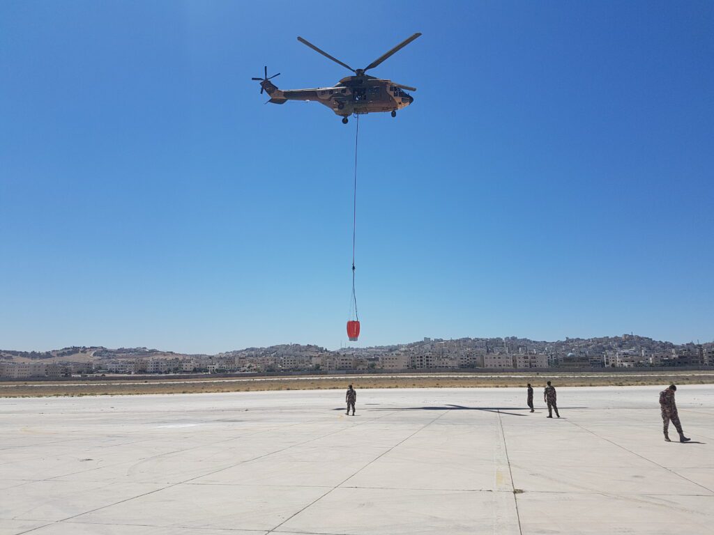 Military heavy-lift helicopter conducting underslung fire bucket hover training over an airfield tarmac