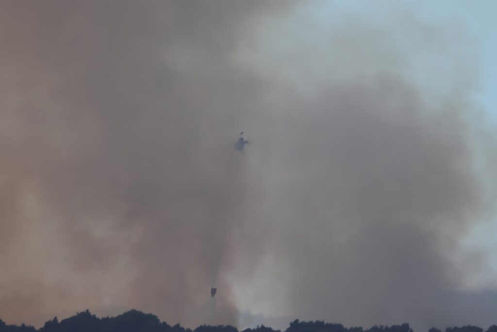 Aerial firefighting helicopter navigating through heavy wildfire smoke while carrying a suspended water bucket.