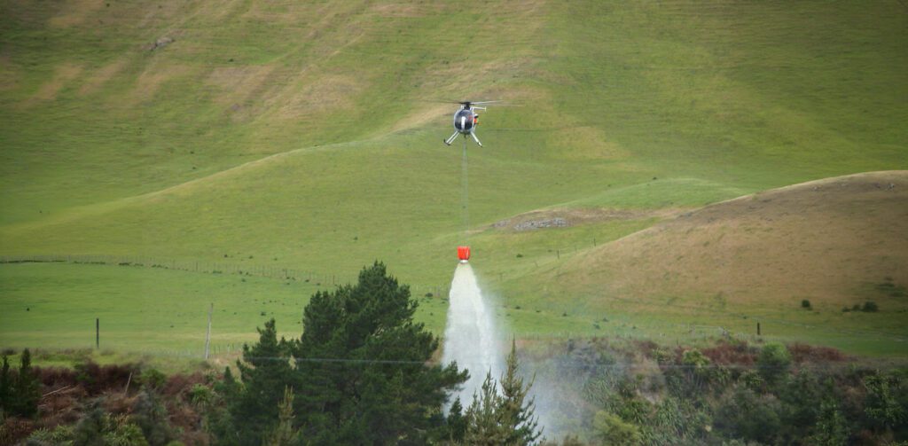 Helicopter performing a precision water drop from an underslung fire bucket over hilly terrain.