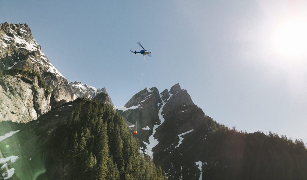 Helicopter equipped with a collapsible fire bucket executing aerial firefighting operations in steep, high-altitude mountainous terrain.