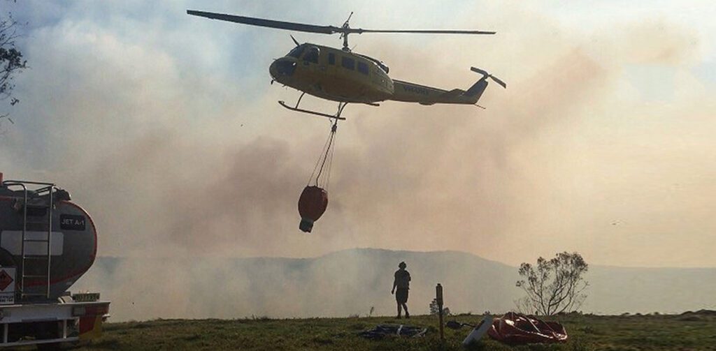 Initial attack firefighting helicopter with an underslung water bucket operating near a smoke-filled fire line.