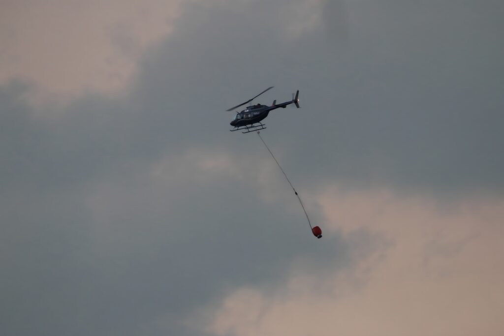 Firefighting helicopter transporting an underslung aerial water bucket on a long-line to a wildfire incident.