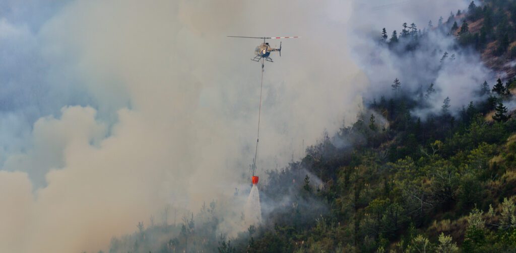 Helicopter navigating heavy smoke to drop water on a steep terrain wildfire using a helibucket.