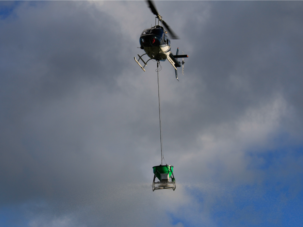 topdressing helicopter flying with a Ground-Effect spreader bucket beneath a cloudy sky