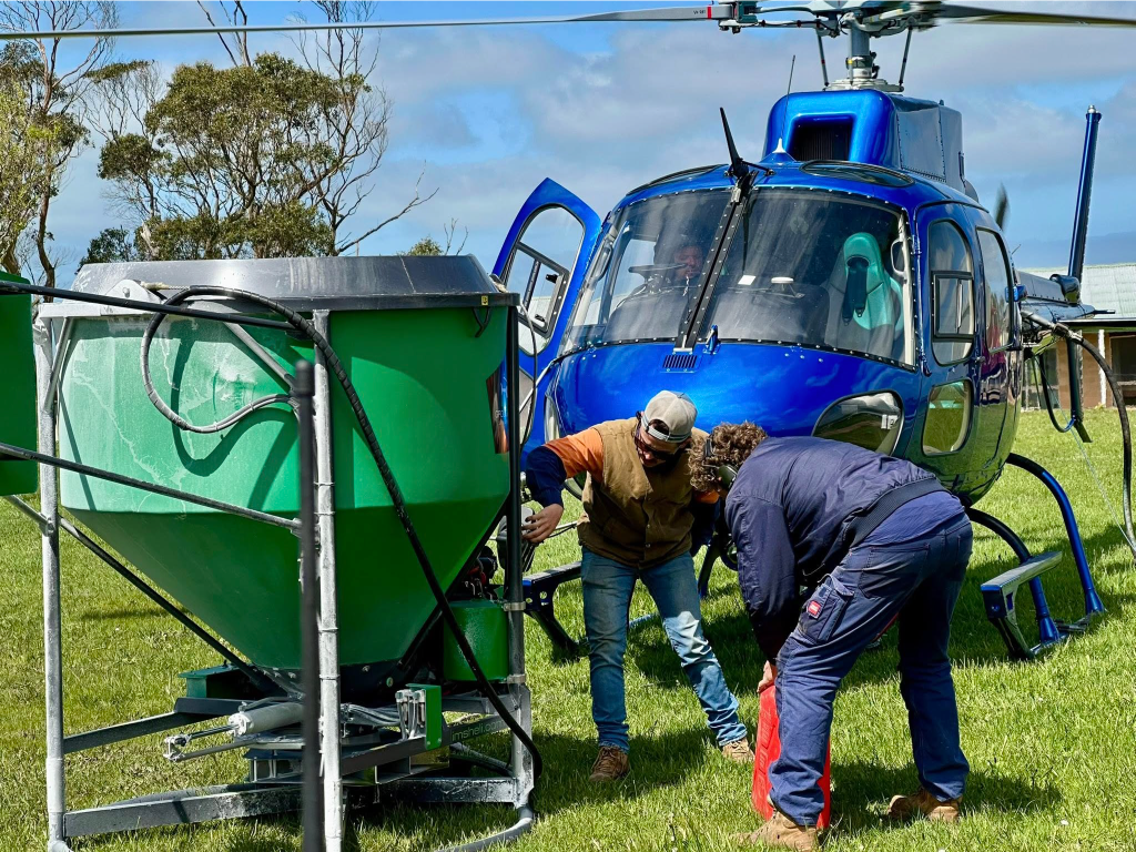 Ground crew inspecting and adjusting a Ground-Effect spreader bucket next to a blue helicopter in a grassy field